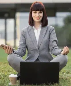 a woman sitting cross legged in the grass with a laptop and a phone